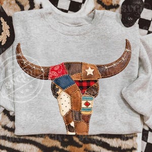 May include: A gray sweatshirt featuring a patchwork bull skull design. The skull is composed of various fabric patches in shades of brown, red, blue, and white, with a star and a bandana print. The horns are brown with white stitching.