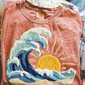 May include: Coral Comfort Colors t-shirt with a textured design of a wave, sun, and rays. The wave is in blue and white, with a yellow sun. The shirt has a crew neck and a tag that says "Comfort Colors".