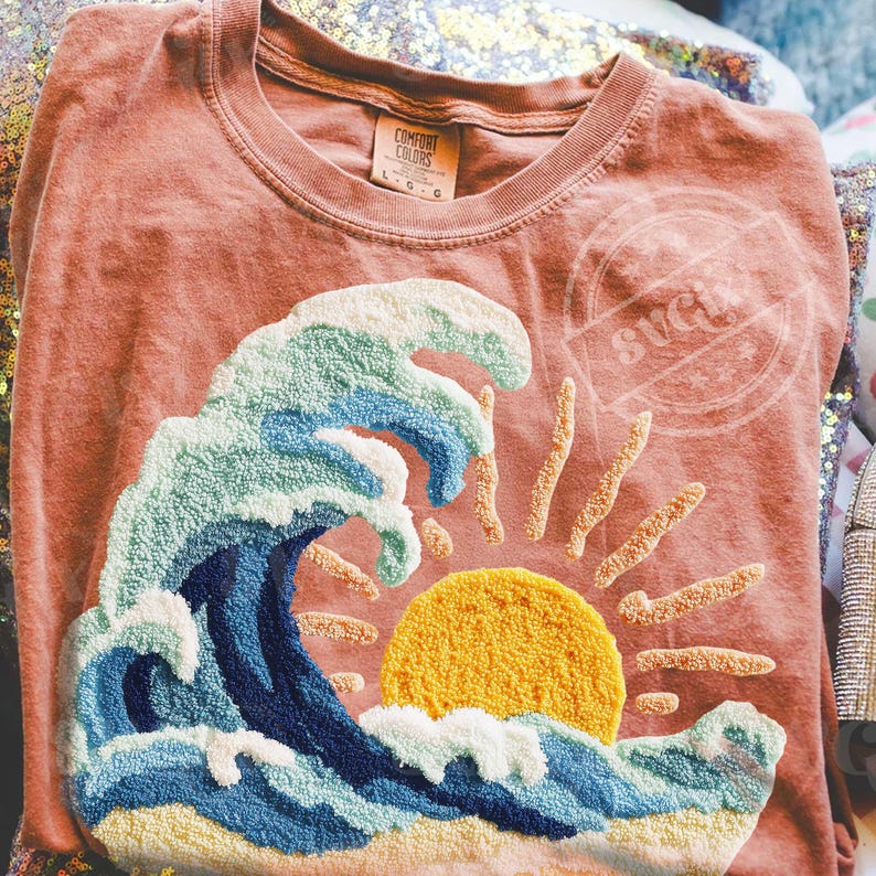May include: Coral Comfort Colors t-shirt with a textured design of a wave, sun, and rays. The wave is in blue and white, with a yellow sun. The shirt has a crew neck and a tag that says "Comfort Colors".