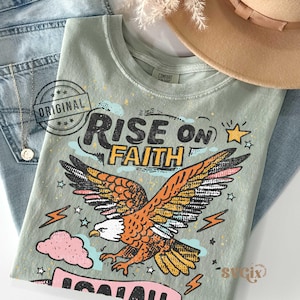 Puede incluir: Una camiseta verde salvia con las palabras "RISE ON FAITH" y una ilustraci&oacute;n de un &aacute;guila con plumas naranjas y amarillas. La camiseta est&aacute; colocada sobre unos vaqueros azules, un collar dorado y un sombrero beige.