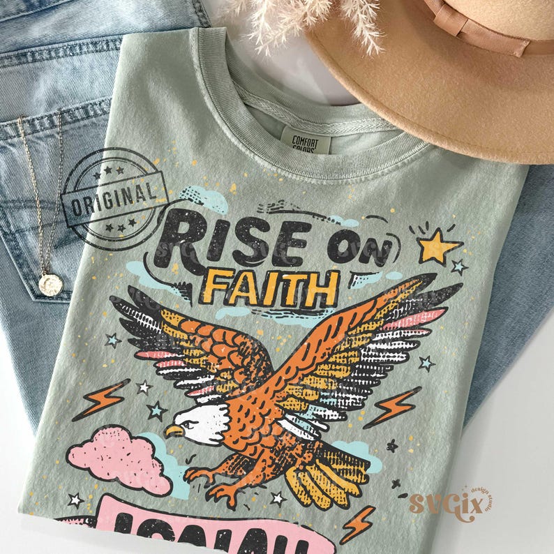 Puede incluir: Una camiseta verde salvia con las palabras "RISE ON FAITH" y una ilustraci&oacute;n de un &aacute;guila con plumas naranjas y amarillas. La camiseta est&aacute; colocada sobre unos vaqueros azules, un collar dorado y un sombrero beige.
