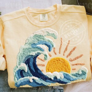 May include: A pale yellow sweatshirt with an embroidered design of a wave, sun, and rays. The wave is in shades of blue, green, and white, with a textured look. The sun is yellow, and the rays are orange.
