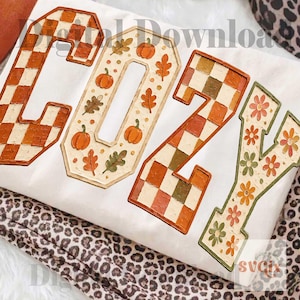 May include: A white fabric surface featuring the word COZY in large block letters. The C has an orange and white checkered pattern, the O features orange pumpkins and leaves, the Z has a checkered pattern, and the Y displays orange and green flowers.