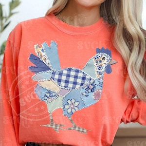 May include: Coral-colored sweatshirt featuring a patchwork chicken design. The chicken is composed of various blue and white patterned fabrics, including gingham and floral prints. The design is centered on the front of the long-sleeved shirt.