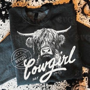 May include: Dark gray sweatshirt featuring a black and white illustration of a Highland cow. The word "Cowgirl" is written in a white script font, with "Say" underneath. A circular logo with stars is visible.
