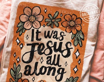 Puede incluir: Una camiseta de color claro con un gráfico rectangular de borde naranja quemado. El diseño incluye ilustraciones florales en tonos melocotón y verde salvia, pequeños detalles de estrellas blancas y el texto negro "It was JESUS all along."