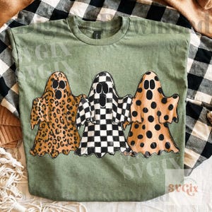 May include: An olive green short-sleeved t-shirt featuring a graphic print of three ghosts. The left ghost has a brown leopard print, the center ghost has a black and white checkerboard pattern, and the right ghost has a brown polka dot pattern.