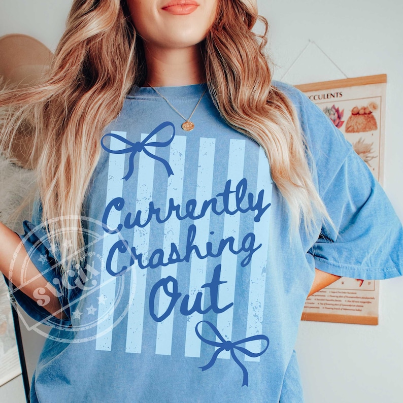 May include: Light blue t-shirt with the text "Currently Crashing Out" in a script font. The design includes light blue vertical stripes and bow accents. The shirt has a relaxed fit and short sleeves.