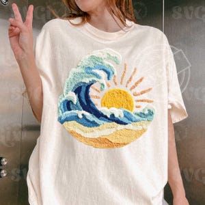 May include: A cream-coloured t-shirt with an embroidered design of a wave, sun, and beach. The wave is in shades of blue and green, the sun is yellow, and the beach is tan. The shirt has short sleeves and a relaxed fit.