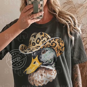 May include: Dark green t-shirt featuring a rhinestone-studded duck wearing a leopard print cowboy hat. The duck has a yellow beak and is embellished with rhinestones. The shirt has a distressed look.