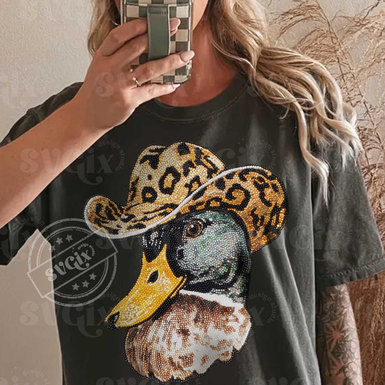 May include: Dark green t-shirt featuring a rhinestone-studded duck wearing a leopard print cowboy hat. The duck has a yellow beak and is embellished with rhinestones. The shirt has a distressed look.