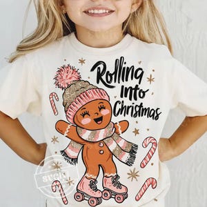 May include: Cream t-shirt with a gingerbread man design. The gingerbread man wears a pink and gold hat, scarf, and roller skates. The text "Rolling Into Christmas" is above the gingerbread man. Candy canes and snowflakes complete the festive design.