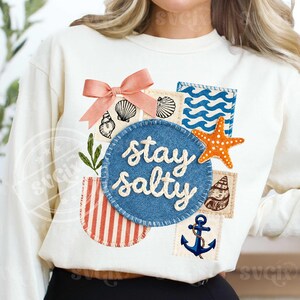 Peut inclure: Sweat-shirt cr&egrave;me avec un collage sur le th&egrave;me de la plage. Comprend un cercle bleu avec le texte "stay salty", des coquillages, une &eacute;toile de mer orange et une ancre bleue. Un n&oelig;ud rose et un vase ray&eacute; ajoutent des d&eacute;tails.