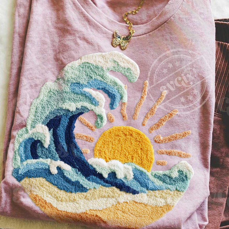 May include: A pale pink t-shirt with an embroidered design of a wave, sun, and beach. The wave is in blue, green, and white, with a yellow sun and sandy beach. A gold butterfly pendant hangs near the neckline. The design evokes a coastal theme.