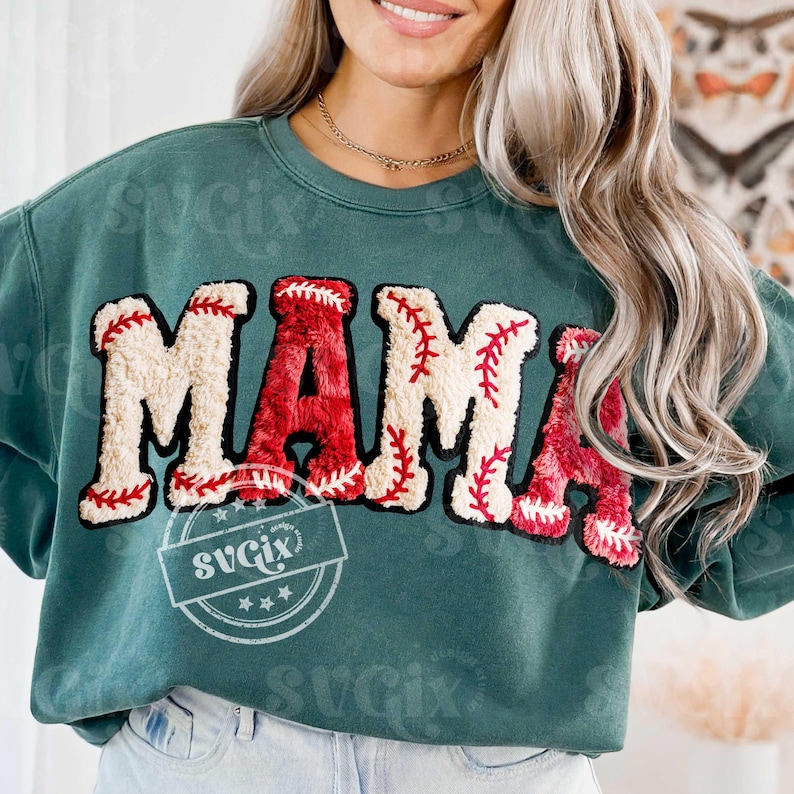 May include: Teal green sweatshirt with the word "MAMA" in a baseball-themed design. The letters are a combination of red and off-white, with red stitching details mimicking a baseball. The sweatshirt has a relaxed fit.
