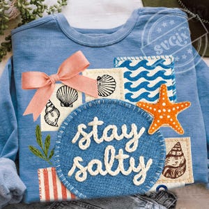 Peut inclure: Sweat-shirt bleu clair orn&eacute; d'un motif d&eacute;coratif avec un n&oelig;ud rose, des coquillages, une &eacute;toile de mer et l'inscription "stay salty". Le motif comprend des patchs avec des motifs de vagues et des illustrations de coquillages.