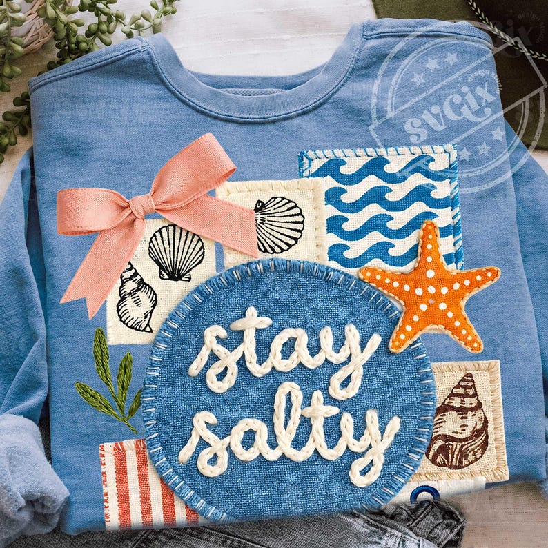 Peut inclure: Sweat-shirt bleu clair orn&eacute; d'un motif d&eacute;coratif avec un n&oelig;ud rose, des coquillages, une &eacute;toile de mer et l'inscription "stay salty". Le motif comprend des patchs avec des motifs de vagues et des illustrations de coquillages.