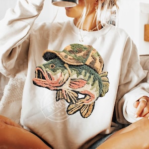 May include: Cream-coloured sweatshirt with an embroidered design of a fish wearing a camouflage hat. The fish is green with pink and brown accents. The design is centered on the front of the sweatshirt, offering a unique, outdoorsy aesthetic.