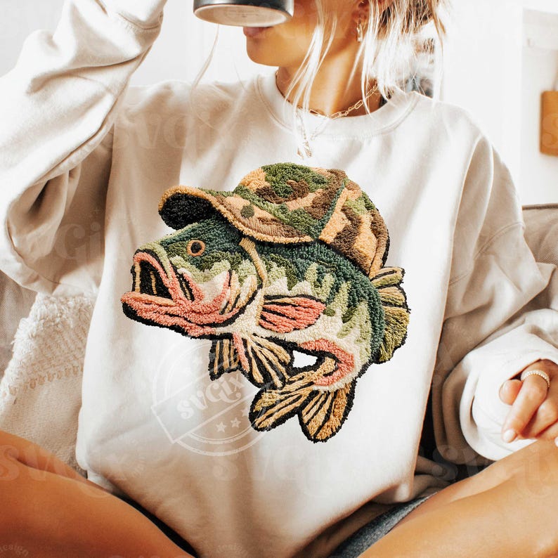 May include: Cream-coloured sweatshirt with an embroidered design of a fish wearing a camouflage hat. The fish is green with pink and brown accents. The design is centered on the front of the sweatshirt, offering a unique, outdoorsy aesthetic.