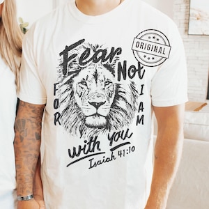 Puede incluir: Camiseta color crema con un detallado rostro de le&oacute;n en blanco y negro. La camiseta tiene las palabras "Fear Not, For I Am With You" e "Isaiah 41:10" impresas, junto con un sello circular "ORIGINAL".