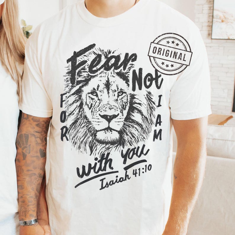 Puede incluir: Camiseta color crema con un detallado rostro de le&oacute;n en blanco y negro. La camiseta tiene las palabras "Fear Not, For I Am With You" e "Isaiah 41:10" impresas, junto con un sello circular "ORIGINAL".