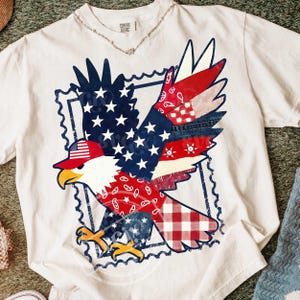 Puede incluir: Camiseta blanca con un diseño de águila patriótica. El águila está representada con las alas extendidas, incorporando elementos de la bandera estadounidense, incluyendo estrellas, rayas y una combinación de colores rojo, blanco y azul. La camiseta tiene el texto "Comfort Colors".