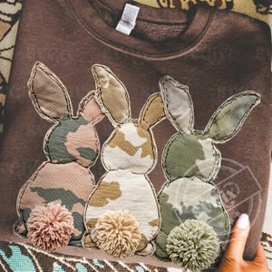 Könnte beinhalten: Ein braunes Sweatshirt mit drei aufgenähten Hasen. Jeder Hase hat ein Camouflagemuster in Rosa-, Beige- und Grüntönen. Die Hasen haben flauschige Pompon-Schwänze und sind auf das Sweatshirt genäht.