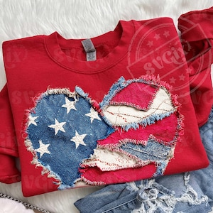 May include: A red sweatshirt featuring a distressed heart design. The heart is made of patches of blue denim with white stars, red fabric, and white lace-like fabric. The sweatshirt is paired with blue denim jeans.