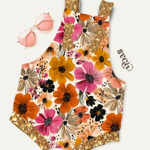 May include: A floral-patterned romper with a cream background, featuring orange, pink, and yellow flowers. The romper has gold glitter accents on the straps and edges. Pink sunglasses rest on the side. The design includes the text "svgix".