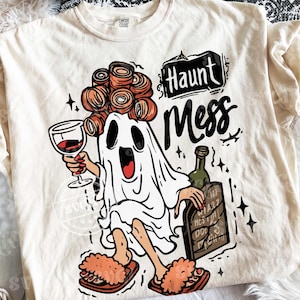May include: Cream-colored t-shirt with a Halloween-themed graphic. The design features a cartoon ghost wearing curlers, holding a wine glass and a bottle of wine. Text on the shirt reads "Haunt Mess."