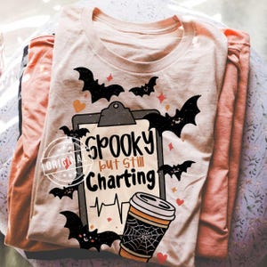 May include: A light peach-colored t-shirt featuring a graphic of a clipboard with the text "Spooky but still Charting" written in black and orange. The design includes black bats, small orange hearts, and a coffee cup with a spiderweb pattern.