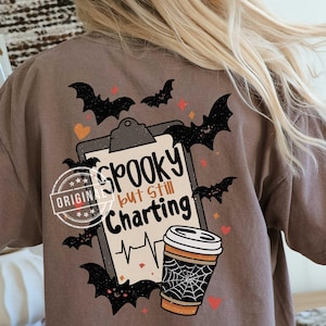 May include: A brown t-shirt featuring a graphic on the back of a clipboard with the text "Spooky but Still Charting" and a heartbeat line. The design includes black bats, orange hearts, and a coffee cup with a spiderweb pattern on the side.