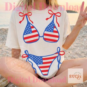 May include: A cream-colored t-shirt featuring a red, white, and blue bikini design. The bikini top and bottom are decorated with the American flag. The top has red bows on the straps, and the bottom has blue ties.