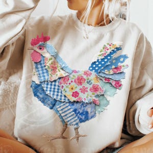 May include: A white sweatshirt featuring a patchwork rooster design. The rooster is made of various blue and floral patterned fabrics, with a pink comb and wattle. The bird's legs are a light brown color.