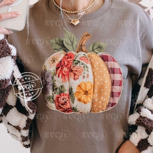 May include: A gray sweatshirt featuring a patchwork pumpkin design. The pumpkin is segmented with floral, polka dot, and plaid patterns in shades of red, orange, and purple. The sweatshirt has a round neckline.
