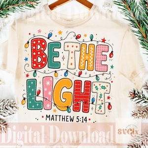 May include: A cream-colored t-shirt featuring the colorful text BE THE LIGHT and MATTHEW 5:14. The letters are decorated with various patterns and colors, surrounded by a string of multi-colored Christmas lights and small star shapes.