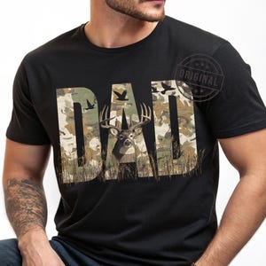May include: A black t-shirt with a camouflage pattern and the word "DAD" in large letters. A deer with antlers is in the center of the word "DAD".