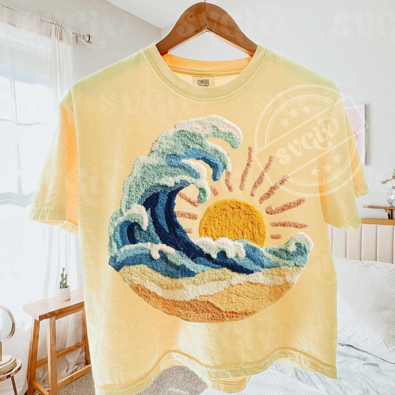 May include: Yellow t-shirt with an embroidered design of a wave, sun, and beach scene. The wave is in blue and green, with a yellow sun and sandy beach. The shirt is hanging on a wooden hanger.
