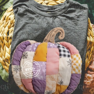 May include: A grey t-shirt with a decorative, patchwork pumpkin. The pumpkin is made of various fabrics in shades of purple, pink, yellow, and white, with black spiderweb accents. The pumpkin has a brown stem.