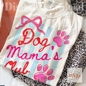 May include: Cream-colored t-shirt with the text "Dog Mama's Club" in red and pink script, accented with a pink bow and paw prints. Light blue vertical stripes add a playful touch to the design. The shirt is paired with leopard print pants.