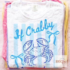 May include: White t-shirt with a blue crab illustration and the words "If Crabby" in a playful font. The design includes blue vertical stripes and bow accents. The shirt is folded on top of other colorful shirts.