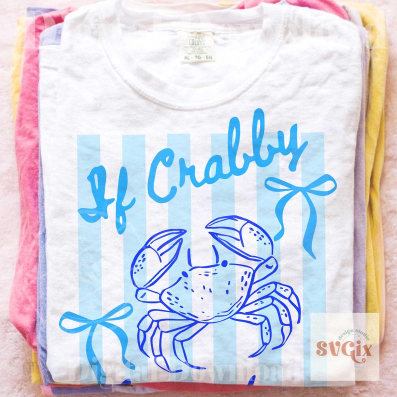 May include: White t-shirt with a blue crab illustration and the words "If Crabby" in a playful font. The design includes blue vertical stripes and bow accents. The shirt is folded on top of other colorful shirts.