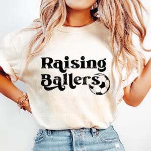 May include: A white t-shirt with black text that reads "Raising Ballers" and a black and white soccer ball graphic.