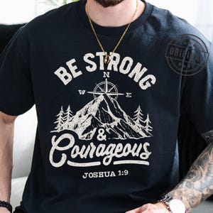 May include: Black t-shirt with a white graphic design. The design features the words "Be Strong & Courageous" above a mountain and compass illustration. The text "Joshua 1:9" is printed below the graphic. A gold chain necklace is visible.