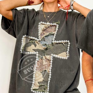 May include: Dark gray t-shirt featuring a large cross design. The cross is made of distressed fabric with a camouflage pattern and is outlined with faux pearls. The shirt has short sleeves and a relaxed fit.
