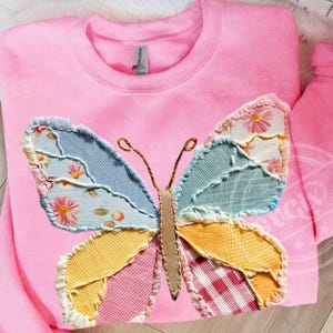 May include: A pink sweatshirt featuring a large, colorful butterfly applique. The butterfly wings are made of patchwork fabric in shades of blue, yellow, pink, and white, with floral and gingham patterns. The butterfly's body is a tan color.