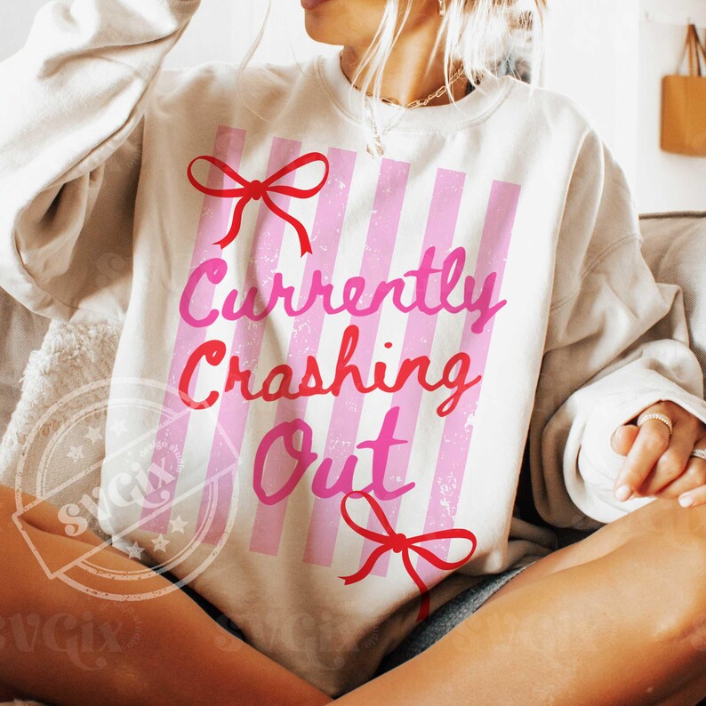 May include: Cream-colored sweatshirt with pink vertical stripes and the text "Currently Crashing Out" in pink and red. The design includes red bow accents. A casual, comfortable top.