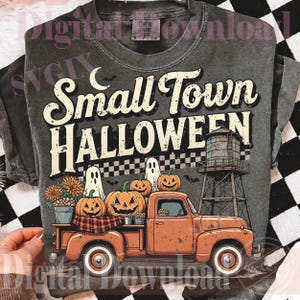May include: A charcoal gray t-shirt featuring a graphic print with the text Small Town HALLOWEEN. The design includes an orange pickup truck carrying pumpkins, two ghosts, a water tower, and a checkered pattern against a dark background.