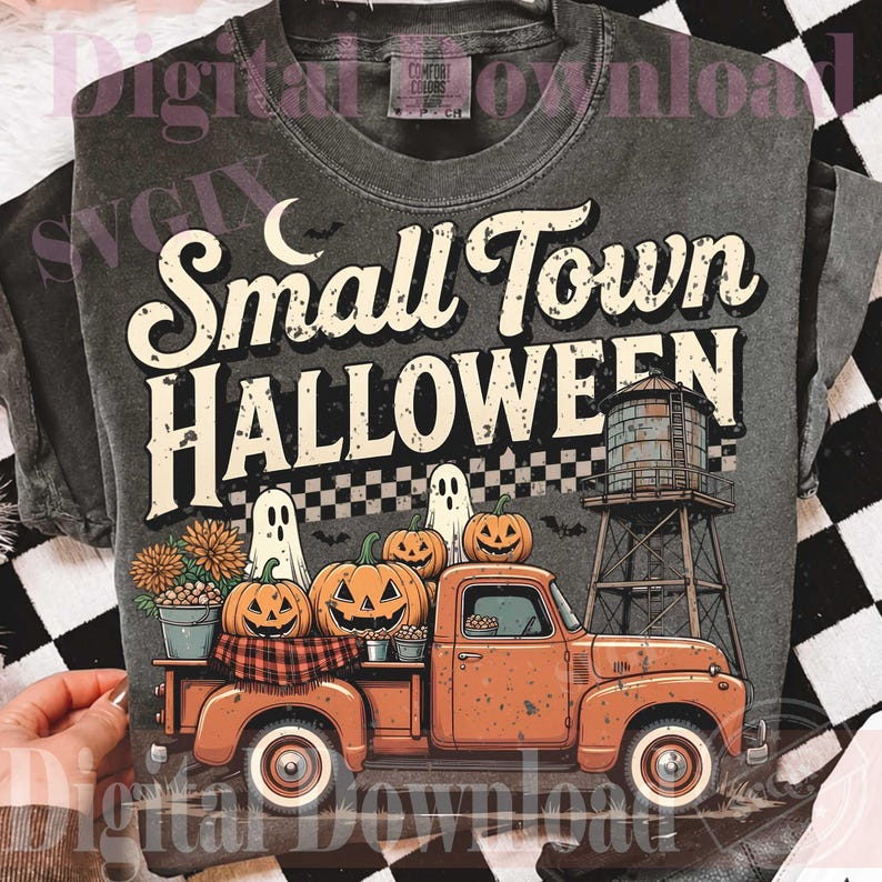 May include: A charcoal gray t-shirt featuring a graphic print with the text Small Town HALLOWEEN. The design includes an orange pickup truck carrying pumpkins, two ghosts, a water tower, and a checkered pattern against a dark background.