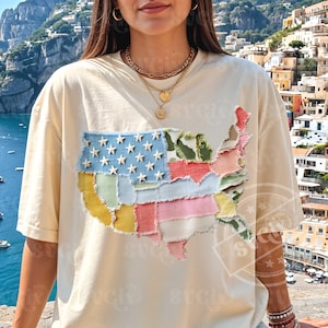 Puede incluir: Camiseta color crema con un diseño de patchwork del mapa de Estados Unidos. El mapa está compuesto por varias piezas de tela en azul con estrellas blancas, rojo, rosa, verde y amarillo. La camiseta tiene un corte holgado y es de estilo casual.